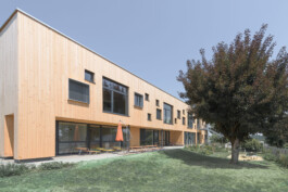 Holzfassade eines Kindergartens und Garten mit Spielgeräten und Bestuhlung 